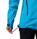 A woman wearing Columbias Womens Saudan Pro™ 3L Shell Jacket in bright blue adjusts the hem drawstring. The waterproof shell features a visible zipper, Velcro cuffs, and is styled with black pants for extreme weather protection.