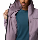 A woman wears a teal shirt and a light purple Columbia Womens Khyex Pro™ Wind Jacket, slightly open to reveal the lining. Only her lower face is visible. This ultralight jacket is wind-resistant and offers UPF 50 sun protection.