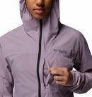 A person wears a light purple Columbia Womens Khyex Pro™ Wind Jacket with the hood up, unzipping a small chest pocket. The Columbia logo appears on the left side of this ultralight, wind-resistant jacket.