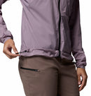 A person adjusts the hem drawcord of a Columbia Womens Khyex Pro™ Wind Jacket in light purple, paired with brown pants with a zippered pocket. Only the torso and part of the arms are shown.