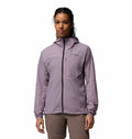 A woman stands facing forward against a white background, wearing the Columbia Womens Khyex Pro™ Wind Jacket in light purple with UPF 50 sun protection and ultralight, wind-resistant design, paired with beige pants.