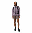A person faces forward, wearing a light purple Columbia Womens Khyex Pro™ Wind Jacket with UPF 50, brown shorts, black hiking shoes, and a black backpack against a white background.