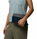 A person in a light blue short-sleeve shirt and olive green pants holds a Columbia Womens Trailborne 2.5L Waterproof Shell Jacket with a small zipper pocket; only their upper body is visible.
