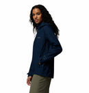 A woman with long curly hair stands sideways, hand in her pocket, wearing Columbia’s Women’s Trailborne 2.5L Waterproof Shell Jacket in navy blue and olive green pants against a plain white background.