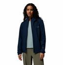 A woman with long, curly dark hair wears a Columbia Womens Trailborne 2.5L Waterproof Shell Jacket over a light blue shirt and olive green pants, standing against a plain white background.