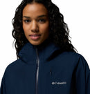 A woman with long, curly hair wears a navy blue Columbia Womens Trailborne 2.5L Waterproof Shell Jacket, zipped to the collar. The Columbia logo appears on her chest against a plain white background.