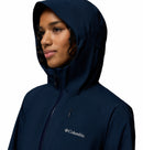 A woman in a dark blue Columbia Womens Trailborne 2.5L Waterproof Shell Jacket, featuring the Columbia logo on the chest, looks to the side against a plain white background.