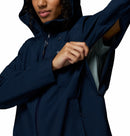 A woman wearing the Columbia Womens Trailborne 2.5L Waterproof Shell Jacket in dark blue unzips an underarm vent, revealing a light inner lining. She is shown from shoulders to waist against a white background.