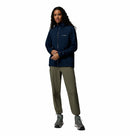 A person with long curly hair wears the Columbia Womens Trailborne 2.5L Waterproof Shell Jacket in navy, olive green pants, and gray sneakers while standing against a plain white background.