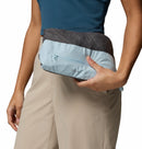 A person in beige pants and a teal top holds the Columbia Womens Trailborne 2.5L Waterproof Shell Jacket, folded into its light blue and gray zippered pouch, against their hip with one hand.