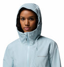 A woman faces forward, looking at the camera, wearing a light blue Columbia Womens Trailborne 2.5L Waterproof Shell Jacket with the hood up and zipped, against a plain white background.