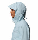A person in profile wears a light blue Columbia Womens Trailborne 2.5L Waterproof Shell Jacket with the hood up and cinched, covering most of the head, set against a plain white background.