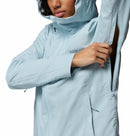 A person wearing a light blue Columbia Womens Trailborne 2.5L Waterproof Shell Jacket unzips an underarm vent. The packable rain jacket features a front zipper and the Columbia logo on the chest. The person’s face is partially visible.