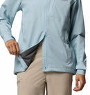 A person wearing a light blue Columbia Womens Trailborne 2.5L Waterproof Shell Jacket and beige pants unzips it to reveal a mesh-lined interior pocket. Their face is not visible in the image.