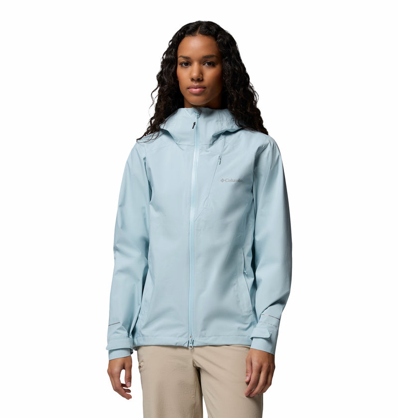 A woman with long curly hair wears the Columbia Womens Trailborne 2.5L Waterproof Shell Jacket in light blue and beige pants, standing facing forward against a plain white background.