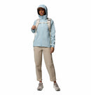 A person wearing a light blue Columbia Womens Trailborne 2.5L Waterproof Shell Jacket, beige pants, gray sneakers, and a beige backpack stands facing forward against a white background.