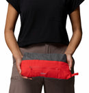 A person in a black shirt and taupe pants holds a red and gray rectangular pouch with front zipper and small handle—ideal for carrying the Columbia Womens Trailborne 2.5L Waterproof Shell Jacket on the go.