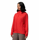 A woman with shoulder-length dark hair and a slight smile stands against a white background, wearing Columbias Womens Trailborne 2.5L Waterproof Shell Jacket in bright red, featuring a high collar and front zippers, paired with taupe pants.