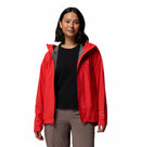 A woman with shoulder-length dark hair wears the Columbia Womens Trailborne 2.5L Waterproof Shell Jacket in bright red over a black shirt and brown pants, standing against a plain white background.