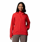 A woman with shoulder-length dark hair wears the Columbia Womens Trailborne 2.5L Waterproof Shell Jacket in bright red and taupe pants, standing with one hand in her pocket against a plain white background.