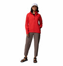 A woman smiles with one hand in her pocket, wearing the Columbia Womens Trailborne 2.5L Waterproof Shell Jacket in red, taupe pants, black sandals, and a white cap against a plain white background.