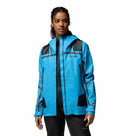 A woman wears the Columbia Womens Whistler Peak Waterproof Shell Jacket in bright blue with black accents and zippers, featuring OutDry Extreme waterproof technology and multiple pockets, standing against a plain white background looking slightly to the side.