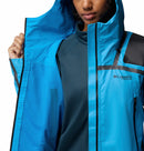 A person partially unzips a bright blue Columbia Womens Whistler Peak Waterproof Shell Jacket, revealing a mesh inner lining and teal base layer. The Columbia logo is visible on the chest of this hiking jacket.