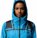 A woman wears a blue Columbia Womens Whistler Peak Waterproof Shell Jacket with black shoulder accents and front zipper, hood up, looking at the camera against a plain white background.