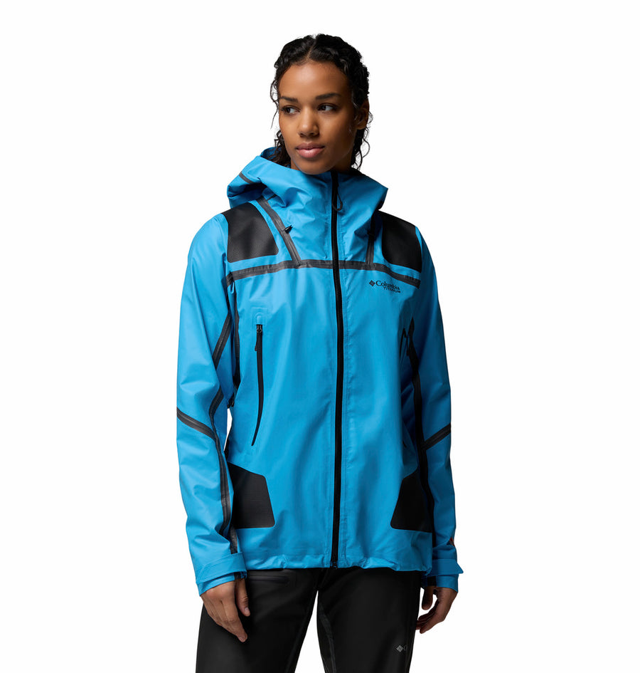 A woman wearing the Columbia Womens Whistler Peak Waterproof Shell Jacket stands against a plain white background. The jacket features a bright blue color, black accents, and zippered pockets.