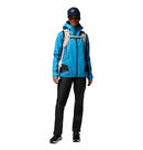A woman stands facing forward in a blue Columbia Womens Whistler Peak Waterproof Shell Jacket, black pants, black sneakers, a blue cap, and a light gray backpack. The outfit is ideal for outdoor adventures. The background is plain white.