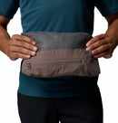 A man in a teal shirt holds the Columbia Mens Trailborne 2.5L Waterproof Shell Jacket, folded in gray and brown, displaying it with both hands against a white background.