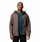 A man faces forward against a plain white background, wearing the Columbia Mens Trailborne 2.5L Waterproof Shell Jacket in brown over a teal shirt and dark pants.