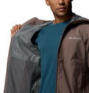 A man in a blue shirt and brown Columbia Mens Trailborne 2.5L Waterproof Shell Jacket opens it to show the patterned lining and mesh pocket. The Columbia logo is on the chest, showcasing its Omni-Tech performance.