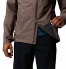 A person wearing Columbia’s Men’s Trailborne 2.5L Waterproof Shell Jacket in brown and black pants stands against a plain white background, holding open a mesh-lined zippered side pocket to reveal the interior.