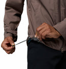 A person adjusts the fit of a Columbia Mens Trailborne 2.5L Waterproof Shell Jacket by tightening the drawstring at the hem; only their torso and hands are shown against a plain background.