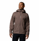 A man wears the Columbia Mens Trailborne 2.5L Waterproof Shell Jacket in brown, featuring a front zipper, standing against a plain white background and smiling while looking slightly to the side.