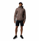 A man stands facing forward against a plain white background, wearing a Columbia Mens Trailborne 2.5L Waterproof Shell Jacket in taupe, black shorts, black sneakers, and a gray cap.