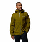 A man wearing the Columbia Mens Trailborne 2.5L Waterproof Shell Jacket in olive green, featuring a hood and Omni-Tech, stands against a plain white background, looking slightly to the side with one hand in his pocket.