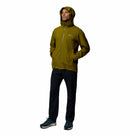 A person stands with hands in pockets, wearing the Columbia Mens Trailborne 2.5L Waterproof Shell Jacket in green, black pants, and sneakers against a white background.