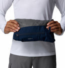 A person in a white long-sleeve shirt and gray pants holds a folded Columbia Mens Trailborne 2.5L Waterproof Shell Jacket, blue and gray, against their torso.
