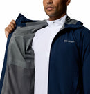 A person wears a navy blue Columbia Mens Trailborne 2.5L Waterproof Shell Jacket over a white zip-up shirt, holding open one side to reveal the patterned gray interior lining.