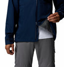 A man wears a Columbia Mens Trailborne 2.5L Waterproof Shell Jacket in navy over a white shirt and gray cargo pants, partially unzipping to show the inner lining. The photo is cropped at the shoulders and knees.