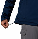 A man in a Columbia Mens Trailborne 2.5L Waterproof Shell Jacket (navy blue) and gray cargo pants tightens the jacket’s hem drawcord with his left hand.