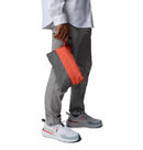 A person in gray pants, a white shirt, and white sneakers stands against a white background, holding a gray and orange zippered pouch while wearing the Columbia Mens Trailborne 2.5L Waterproof Shell Jacket.