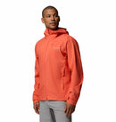 A man wears a Columbia Mens Trailborne 2.5L Waterproof Shell Jacket in bright orange and light gray pants, standing against a plain white background and looking slightly to his left.