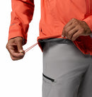 A person wearing a coral Columbia Mens Trailborne 2.5L Waterproof Shell Jacket and light gray pants adjusts the jackets hem drawcord, with only their torso and hands visible.