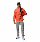 A man wearing a blue cap, Columbia Mens Trailborne 2.5L Waterproof Shell Jacket in orange, gray pants, and white sneakers stands facing forward, holding a backpack with a black strap over his shoulder.