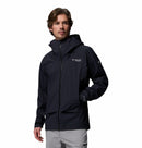 A man in a black Columbia Mens Saudan Pro 3L Waterproof Shell Jacket with a hood, light brown hair, and light gray pants stands against a plain white background, one hand in his pocket.
