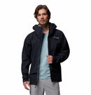 A man wears the Columbia Mens Saudan Pro 3L Waterproof Shell Jacket in black over a light blue shirt and light gray pants, standing against a plain white background and looking forward with a neutral expression.