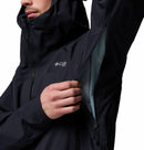 A person in a dark Columbia Mens Saudan Pro 3L Waterproof Shell Jacket with a partially unzipped hood reveals the inner lining and chest logo, while unzipping an underarm ventilation opening.